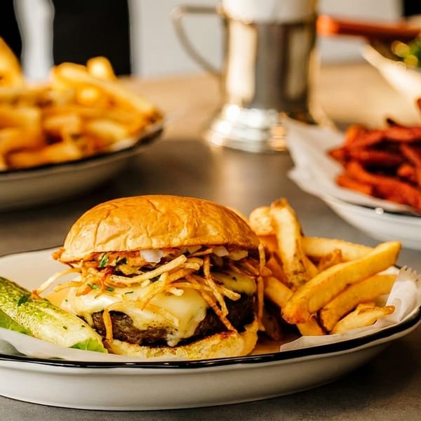 Cheeseburger with fries, lettuce, and pickle on a white plate, with other dishes blurred in the background.